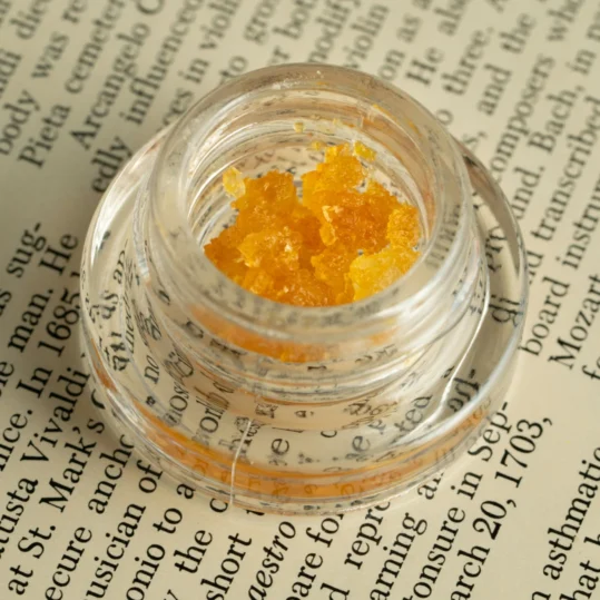 THCA concentrate in a small, clear concentrate jar, sitting on top of a newspaper. The golden, glossy texture of the concentrate contrasts with the printed text and images on the newspaper, highlighting the product's quality and purity in an everyday setting.