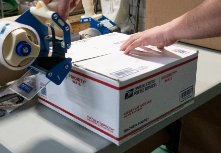 Hands sealing a USPS package with adhesive tape, preparing it for shipment.